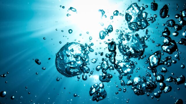 Dark bubbles rising in blue water towards a surface light
